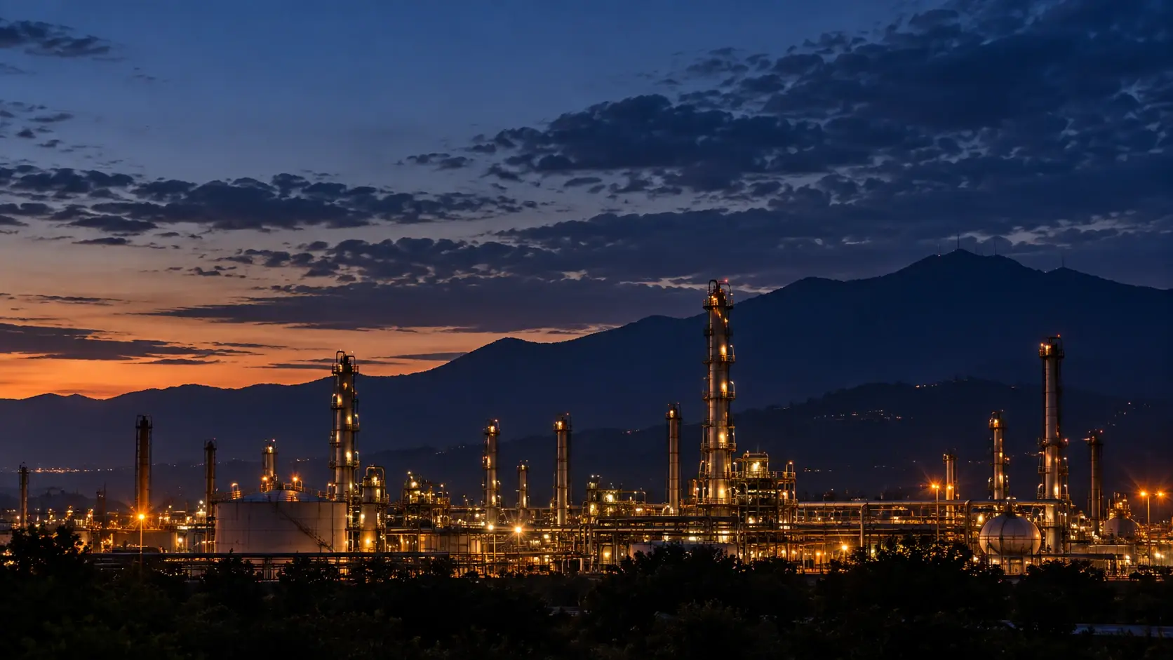 Vista amplia de una refinería iluminada al anochecer, con torres de proceso y la cordillera en silueta al fondo